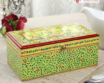 May include: A yellow wooden box with a green and red floral design. The box has a brass handle and a red trim around the top edge. The box is sitting on a brown surface.