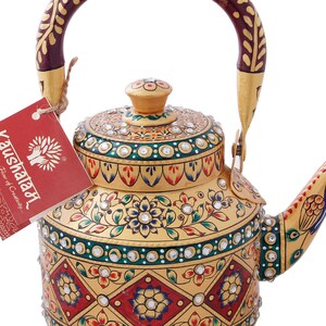 May include: A gold-colored metal teapot with a red and gold handle. The teapot is decorated with intricate floral patterns and rhinestones. The teapot is labeled "Kaushaala,  Flow of Creativity."