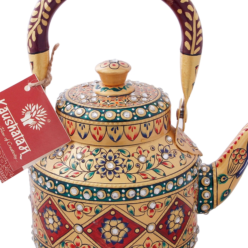 May include: A gold-colored metal teapot with a red and gold handle. The teapot is decorated with intricate floral patterns and rhinestones. The teapot is labeled "Kaushaala,  Flow of Creativity."