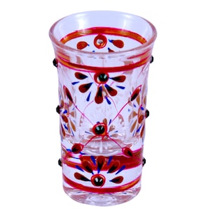 May include: A clear glass shot glass featuring hand-painted floral motifs in red, blue, and gold. The design includes horizontal bands near the rim and base, with small raised black dots applied to the exterior surface around the middle section.