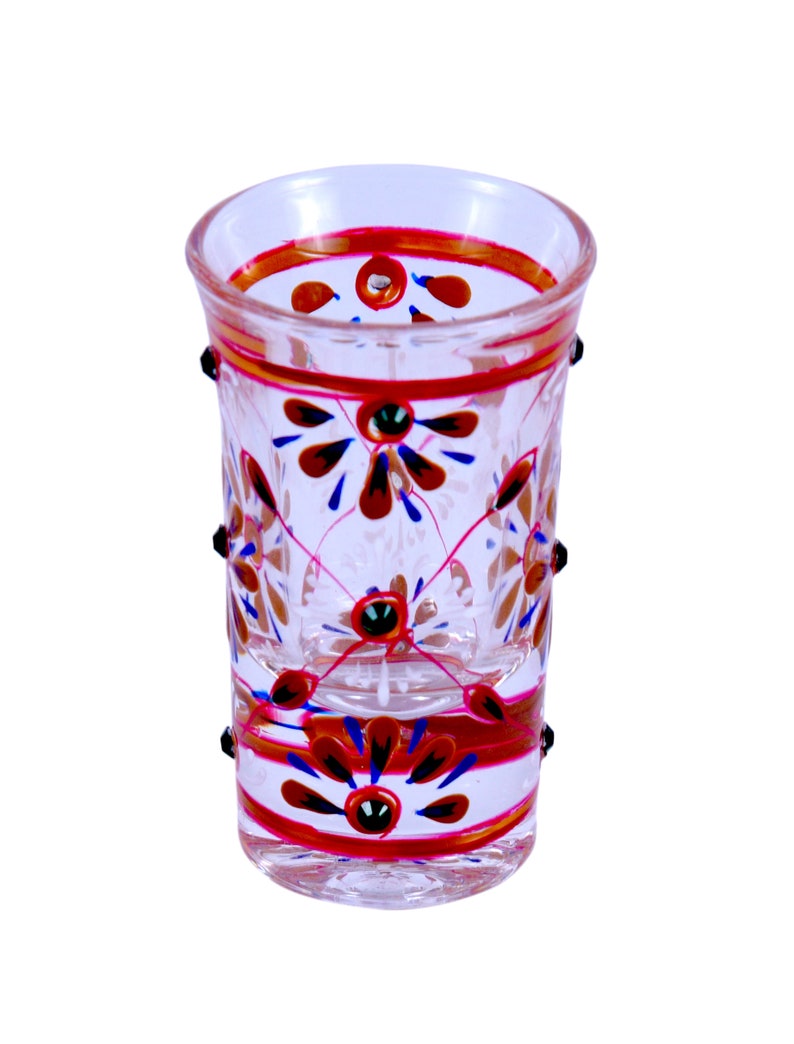 May include: A clear glass shot glass featuring hand-painted floral motifs in red, blue, and gold. The design includes horizontal bands near the rim and base, with small raised black dots applied to the exterior surface around the middle section.