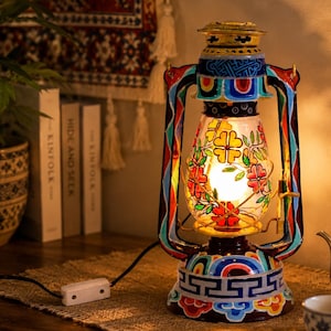 Op de afbeelding: Een decoratieve metalen lantaarnlamp met een handgeschilderd frame in levendige blauwe, rode en gele geometrische patronen. De heldere glazen bol is versierd met kleurrijke bloemmotieven en bevat een gloeiende lamp. Een zwart snoer is bevestigd.