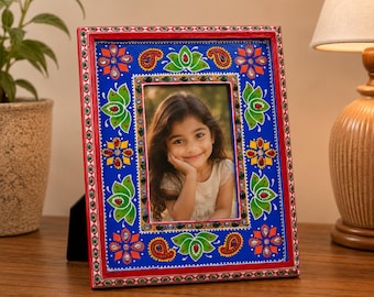 Op de afbeelding: Een rechthoekige fotolijst met een levendige blauwe achtergrond, voorzien van handgeschilderde groene lotusbloemen, oranje paisley-motieven en kleurrijke bloemmotieven. De lijst is rood omrand en versierd met kleine, reflecterende edelstenen.