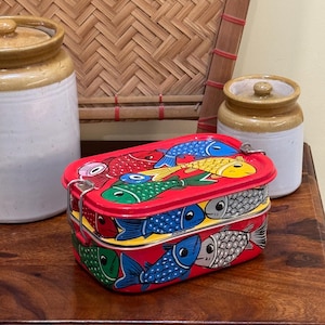 Peut inclure: Une boîte à lunch en métal rouge avec un couvercle à charnière, décorée d'illustrations de poissons colorés. La boîte à lunch est rectangulaire et possède un fermoir argenté. Deux pots en céramique et un tapis en bambou tressé sont en arrière-plan.