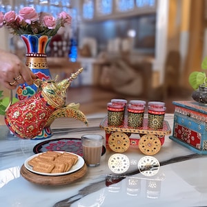 Puede incluir: Una cafetera ornamentada, roja y dorada, con un pico largo, junto a un plato de galletas y un vaso de té. Frascos decorativos están sobre un pequeño carro con ruedas. También hay un jarrón de rosas rosas y una caja pintada.
