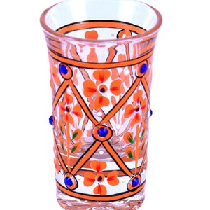 May include: A clear glass tumbler featuring a hand-painted orange lattice pattern with orange floral motifs and small green leaves. The design is accented with small, round, dark blue decorative studs placed at the intersections of the lattice.