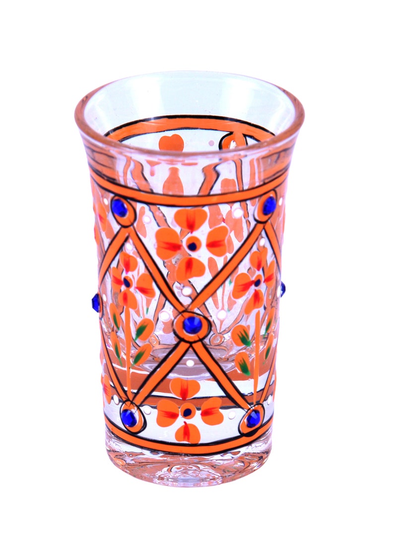 May include: A clear glass tumbler featuring a hand-painted orange lattice pattern with orange floral motifs and small green leaves. The design is accented with small, round, dark blue decorative studs placed at the intersections of the lattice.