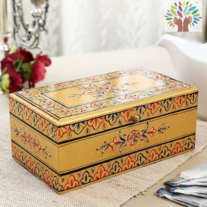 May include: A rectangular, golden-colored wooden tea box with intricate floral designs in red, blue, and pink. The box features a brass knob and is decorated with hand-painted patterns. The tea box is placed on a beige surface.