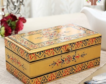 May include: A rectangular, golden-colored wooden tea box with intricate floral designs in red, blue, and pink. The box features a brass knob and is decorated with hand-painted patterns. The tea box is placed on a beige surface.