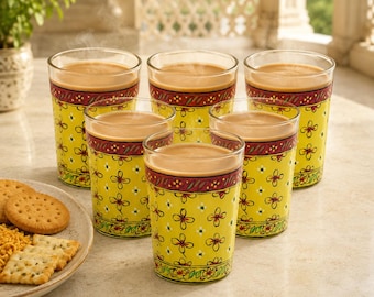 Op de afbeelding: Een set van zes heldere glazen gevuld met een lichtbruine drank. Elk glas heeft een felgele basis versierd met een herhalend patroon van kleine rode en oranje bloemen, geaccentueerd door een horizontale band in donkerrood en groen.