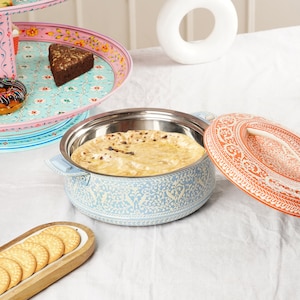 Puede incluir: Un calientaplatos azul claro y acero inoxidable con un diseño floral, que contiene un pan plano. La tapa es naranja con un estampado floral blanco. También se ve una bandeja de servicio escalonada y una bandeja de madera con galletas.