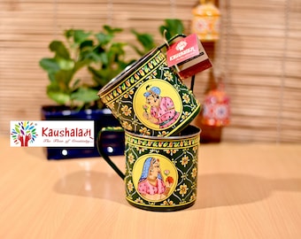 以下が含まれることがあります： 取っ手付きの緑色の金属製のマグカップ2つ。カラフルな花柄と、伝統的なインドの衣装を着た人々の絵が描かれています。マグカップは重ねられています。