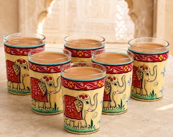 Op de afbeelding: Een set van zes heldere glazen gevuld met een lichtbruine drank. Elk glas is voorzien van een handgeschilderd ontwerp van een beige olifant met een rood en goudkleurig kleed, geaccentueerd door een rode rand met witte bloemen en groen gras.