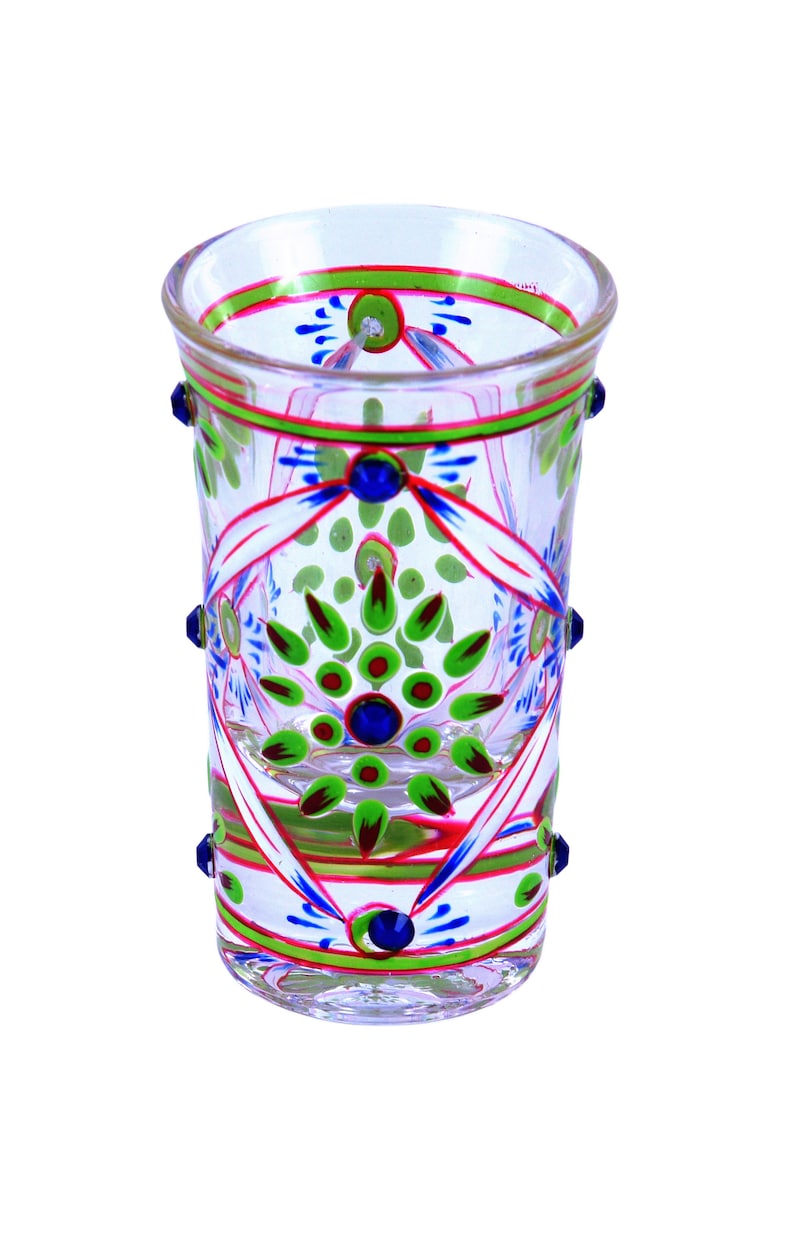 May include: A clear glass tumbler featuring hand-painted floral patterns in vibrant green, blue, and red. The design includes symmetrical petal motifs, thin horizontal bands around the rim and base, and small blue glass studs applied to the exterior.