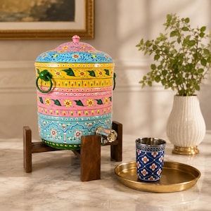 May include: A hand-painted, colorful water dispenser with a floral design, set on a wooden stand. The dispenser has a spigot and green handles. A blue floral patterned cup sits on a gold-colored tray.