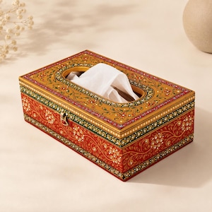 Puede incluir: Un soporte rectangular de madera para caja de pañuelos con una base roja pintada a mano, decorada con complejos patrones florales dorados y pequeños puntos blancos. La tapa color mostaza tiene una abertura ovalada con un pañuelo blanco.