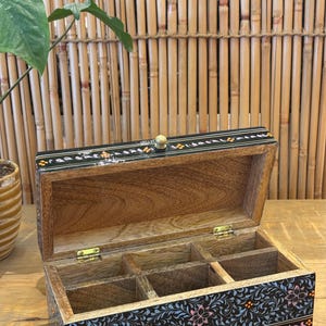 May include: A black wooden box with a floral design and a small wooden knob on the lid. The box has four compartments inside.