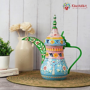 May include: A hand-painted metal teapot with a green handle and spout. The teapot is decorated with floral patterns in pink, blue, yellow, and green. The teapot is sitting on a brown woven mat.