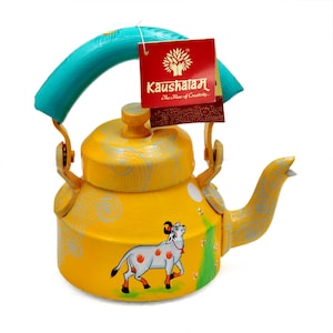 Könnte beinhalten: Gelbe Metall-Teekanne mit blauem Griff und einem bemalten Kuh-Motiv. Die Kanne hat einen weißen Hintergrund mit blauen und gelben Wirbeln. Der Text "Kaushalam The Home of Creativity..." befindet sich auf einem roten Etikett, das am Griff befestigt ist.