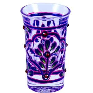 May include: A clear glass shot glass featuring hand-painted floral patterns in vibrant shades of blue, pink, and green. The exterior surface is decorated with raised, rounded red accents arranged in a symmetrical design around the glass body.