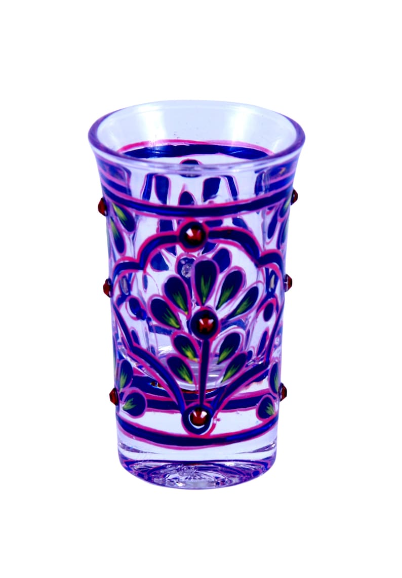 May include: A clear glass shot glass featuring hand-painted floral patterns in vibrant shades of blue, pink, and green. The exterior surface is decorated with raised, rounded red accents arranged in a symmetrical design around the glass body.
