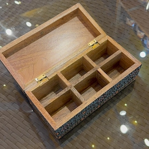 May include: An open wooden box with six compartments. The box is made of light brown wood and has a dark blue and black floral design on the bottom. The box is open and the compartments are empty.