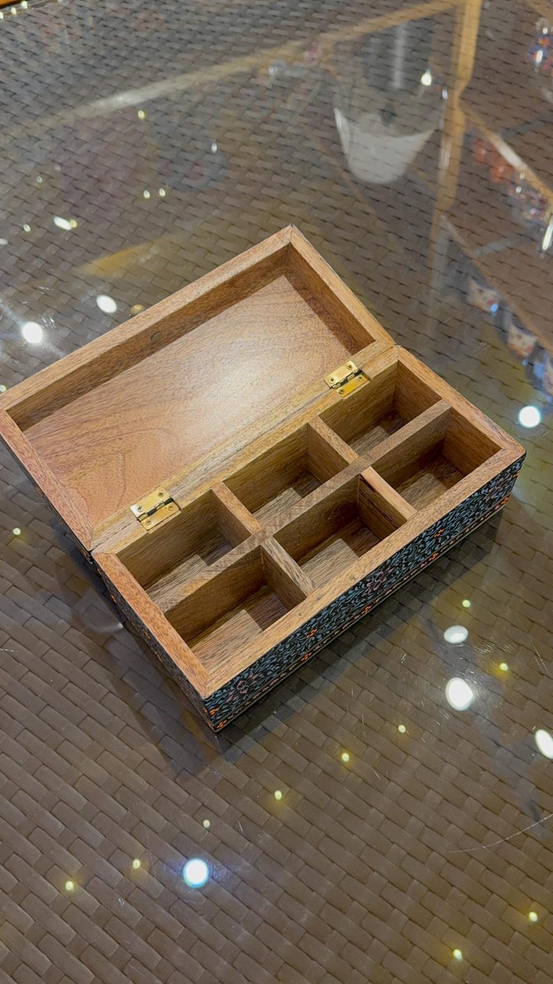 May include: An open wooden box with six compartments. The box is made of light brown wood and has a dark blue and black floral design on the bottom. The box is open and the compartments are empty.
