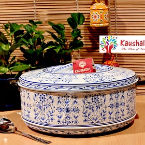 Puede incluir: Un recipiente de almacenamiento de alimentos de cerámica blanco y azul con un diseño floral. El recipiente tiene una tapa con asa y está hecho de metal. El recipiente está sobre una superficie de madera.