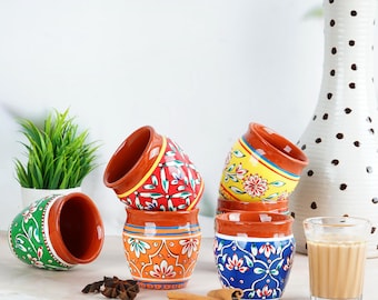 May include: A collection of hand-painted ceramic tea cups in various colors, including red, orange, yellow, green, and blue. Each cup features floral and geometric designs. A glass of tea and spices are also present.