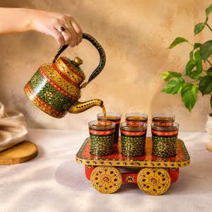 May include: A hand-painted metal teapot with intricate green, red, and gold floral patterns pouring tea into six matching glass tumblers. The glasses sit on a rectangular wooden serving cart with four decorative wheels and floral motifs.