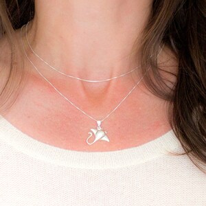 Può includere: Collana con pendente a forma di manta in argento. Il pendente è una rappresentazione stilizzata e dettagliata di una manta. La collana ha una catenina sottile ed è indossata su un maglione color crema.