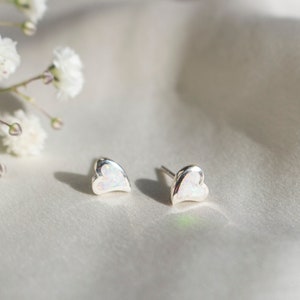 May include: A pair of silver heart-shaped stud earrings with a white, opal-like centre. The earrings are displayed on a soft, white surface, with white flowers in the background. The earrings are small and delicate.