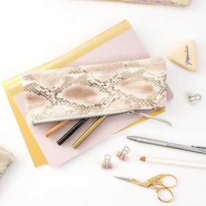 Può includere: Un astuccio con motivo a pelle di serpente con cerniera, appoggiato su un blocco note rosa e oro. All'interno ci sono penne dorate, nere e beige. Sono presenti altri articoli di cancelleria.