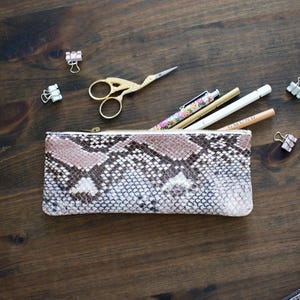 Op de afbeelding: Een etui met slangenpatroon en een gouden rits, open om pennen en potloden te laten zien. Gouden scharen en paperclips liggen verspreid op een donker houten oppervlak. De etui is in bruin-, beige- en wittinten.