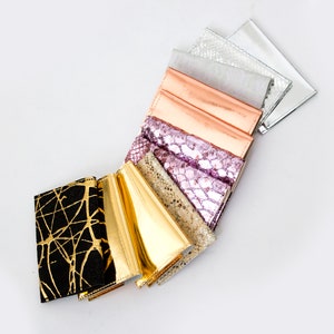 May include: A collection of wallets in various colors and textures, including gold, black with gold accents, metallic silver, and pink. The wallets are arranged in a fan shape against a white background.