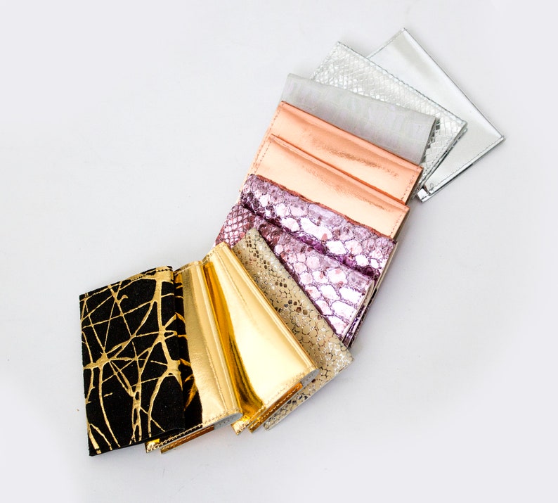 May include: A collection of wallets in various colors and textures, including gold, black with gold accents, metallic silver, and pink. The wallets are arranged in a fan shape against a white background.