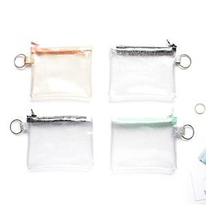 May include: Four small, clear glittery pouches with different colored zippers and key rings. The zipper colors are rose gold, silver, black, and mint green. The pouches are square-shaped and appear to be made of a clear, flexible material.
