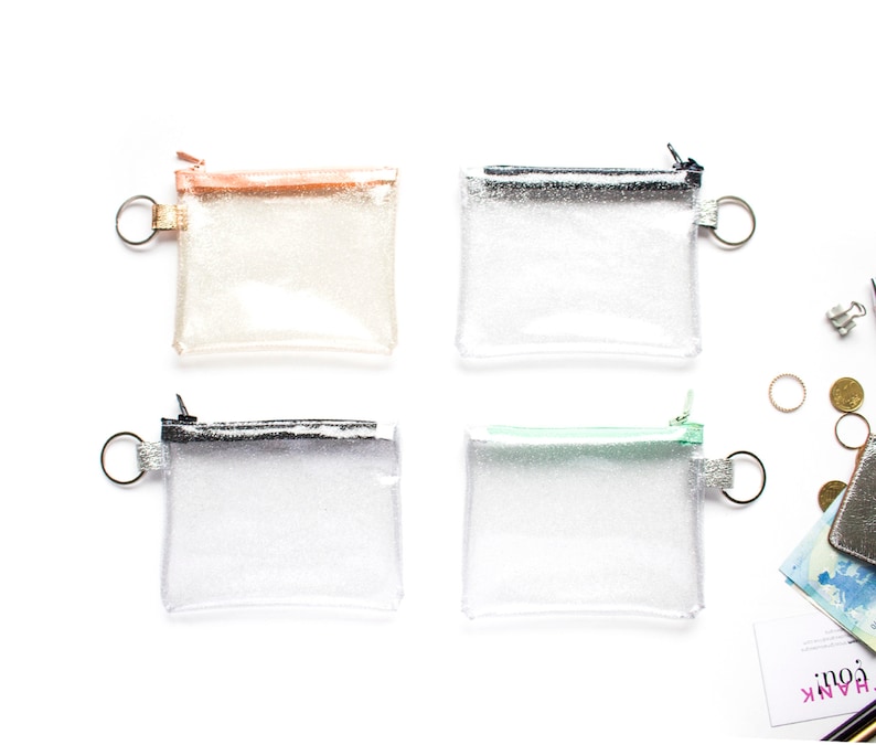 May include: Four small, clear glittery pouches with different colored zippers and key rings. The zipper colors are rose gold, silver, black, and mint green. The pouches are square-shaped and appear to be made of a clear, flexible material.