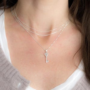 May include: A silver necklace featuring a key pendant. The pendant has a heart-shaped top and intricate detailing. The necklace is layered with three fine chains. The key pendant is approximately 2.5 cm in length.