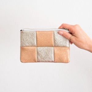 May include: A small, rectangular clutch with a zipper closure. The clutch is divided into a grid pattern of alternating squares of metallic silver and peach-colored leather. The zipper is silver, and the interior is white.