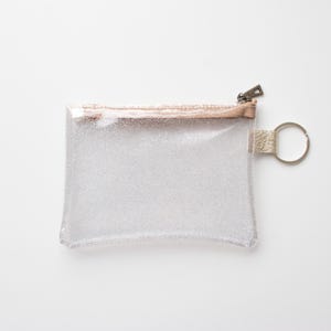 May include: A clear, rectangular glittery pouch with a metallic rose gold zipper and trim. The pouch has a small loop with a silver key ring attached. The background is white.