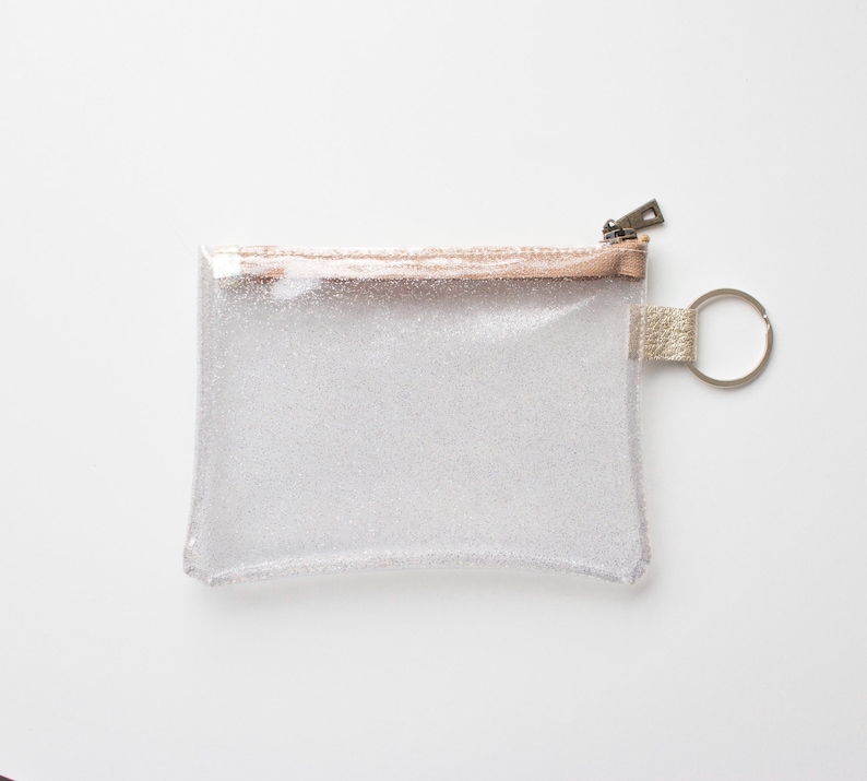 May include: A clear, rectangular glittery pouch with a metallic rose gold zipper and trim. The pouch has a small loop with a silver key ring attached. The background is white.