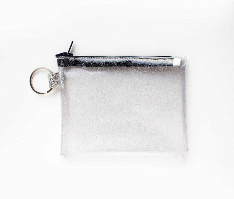 May include: A clear, rectangular glittery pouch with a black zipper and a silver key ring. The pouch is filled with silver glitter and has a black border along the top. The key ring has a small, decorative silver band.