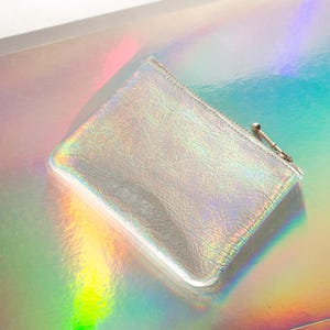 May include: A small, rectangular iridescent coin purse with a zipper closure and a small metal pull. The purse has a textured surface and reflects a rainbow of colors. The background is a similar iridescent material.
