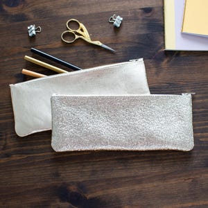 May include: Two metallic silver pencil cases with zippers are displayed on a dark wood surface. One case has a smooth finish, while the other has a textured, crackled appearance. Gold scissors, pencils, and binder clips are also visible.
