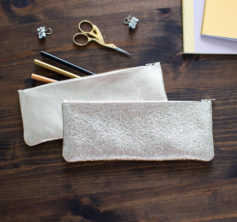 May include: Two metallic silver pencil cases with zippers are displayed on a dark wood surface. One case has a smooth finish, while the other has a textured, crackled appearance. Gold scissors, pencils, and binder clips are also visible.