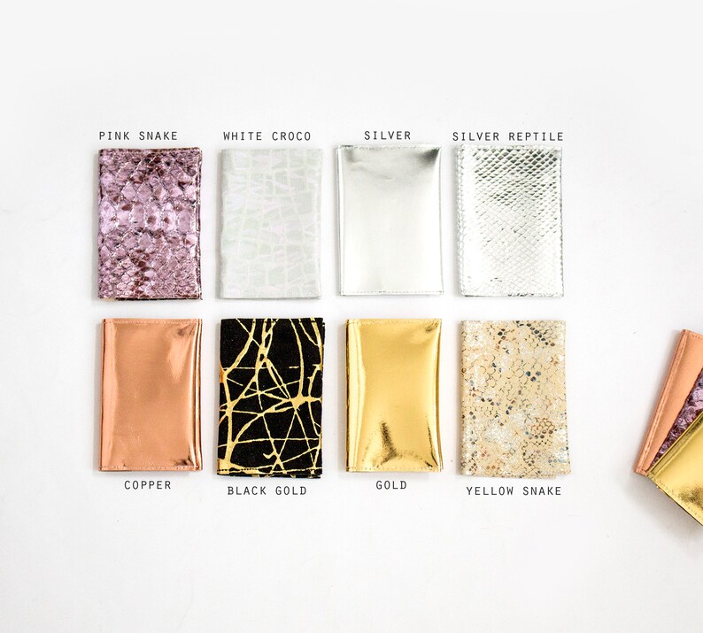 May include: A collection of eight rectangular wallets in various colors and textures. The wallets include pink snake, white croco, silver, silver reptile, copper, black gold, gold, and yellow snake. Each wallet is labeled with its respective color or pattern.