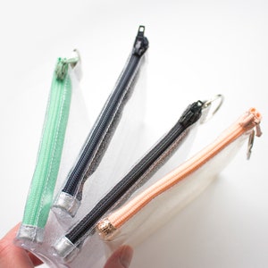 May include: Four clear, rectangular pencil cases with colorful zippers. The zippers are mint green, navy blue, black, and peach. Each case has a silver-colored metal zipper pull. The cases are held in a hand.