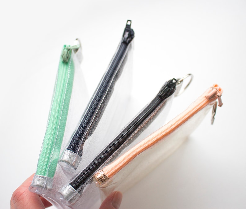 May include: Four clear, rectangular pencil cases with colorful zippers. The zippers are mint green, navy blue, black, and peach. Each case has a silver-colored metal zipper pull. The cases are held in a hand.