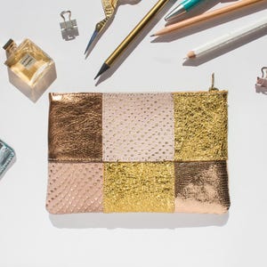 Op de afbeelding: Een rechthoekige clutch met een patchwork-ontwerp in goud, brons en roze tinten. De clutch heeft een ritssluiting en ligt op een wit oppervlak, omringd door briefpapier en een parfumflesje.